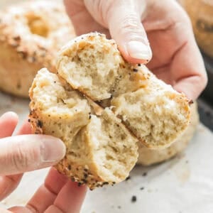 Hands breaking apart a freshly baked 3-ingredient bagel topped with seeds. The interior appears soft and fluffy, while the crust is golden brown and slightly crispy, with visible poppy and sesame seeds scattered on the surface.